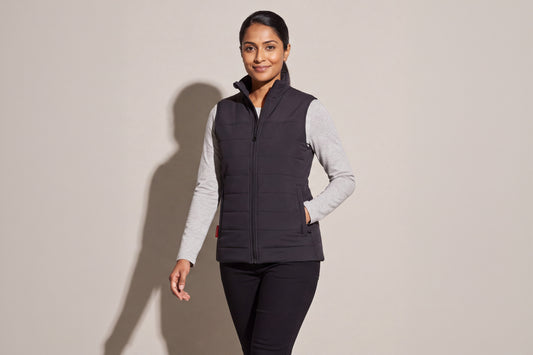 A woman wears a black heated puffer vest over a grey long-sleeve top and black trousers, against a warm grey studio wall, women’s frame