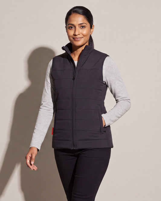 Une femme porte une veste chauffante matelassée noire par-dessus un chandail gris à manches longues et un pantalon noir, devant un mur de studio gris chaud.