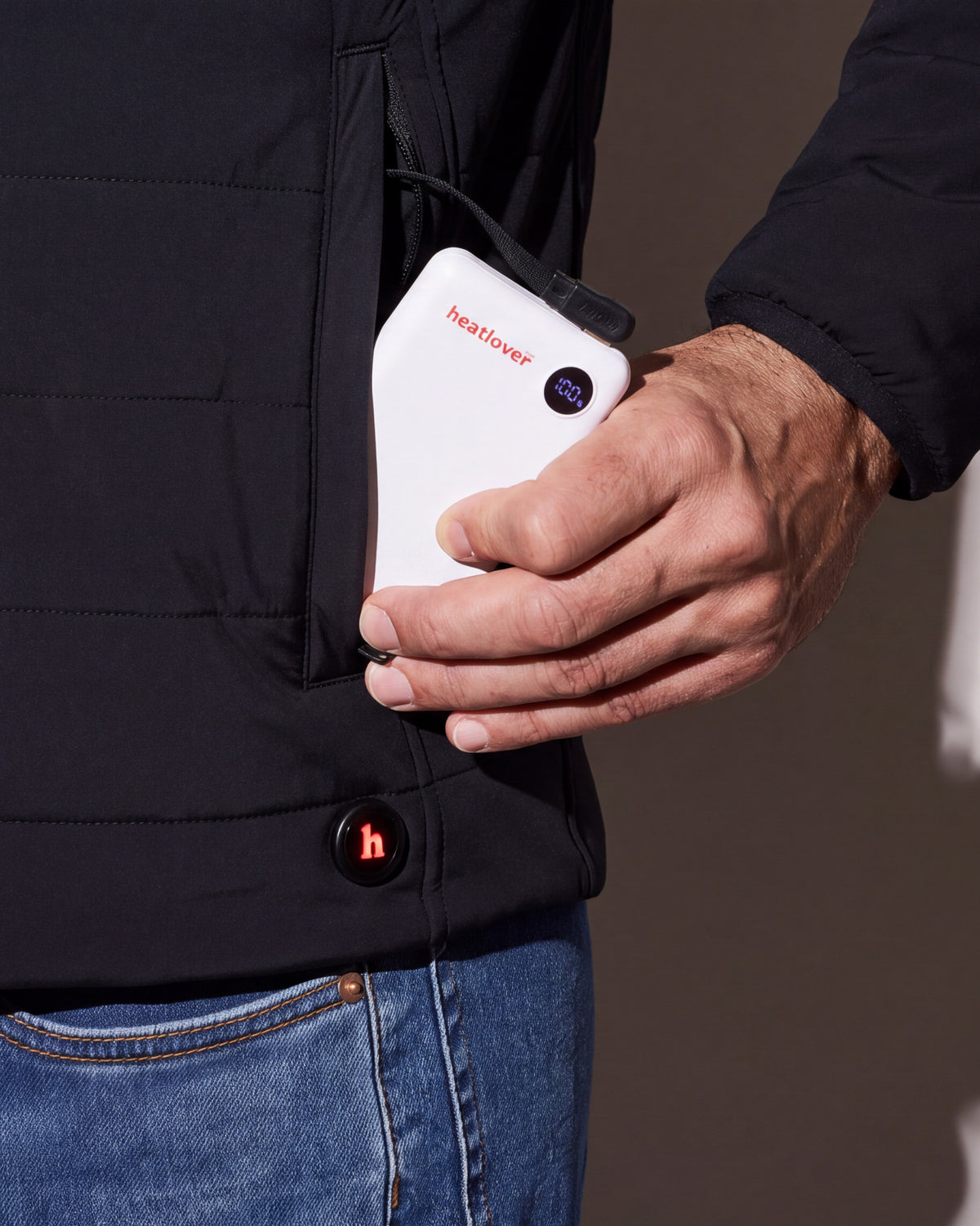 A hand slides the white Heatlover 10K USB-C battery, its charge display lit, into the pocket of a black heated jacket beside the h button.