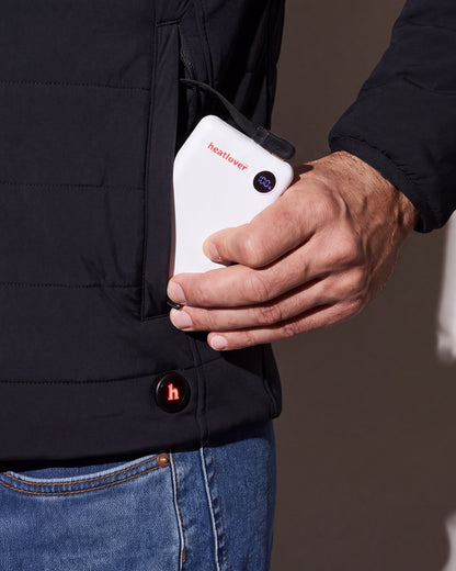 A hand slides the white Heatlover 10K USB-C battery, its charge display lit, into the pocket of a black heated jacket beside the h button.