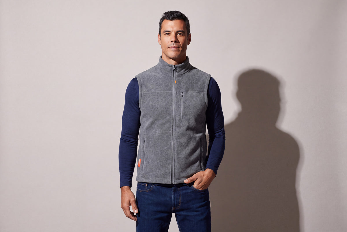 A man wears a grey heated fleece vest zipped over a navy long-sleeve top and jeans, one hand in the pocket, against a warm grey studio wall.