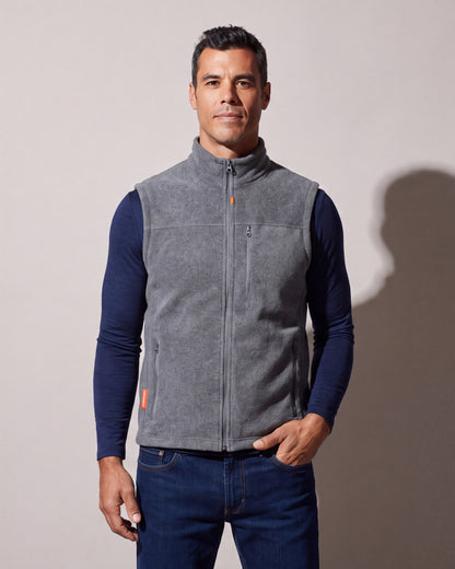 A man wears a grey heated fleece vest zipped over a navy long-sleeve top and jeans, one hand in the pocket, against a warm grey studio wall.