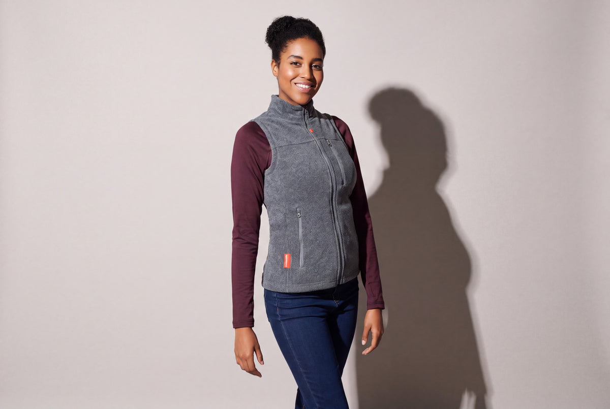 A woman wears a grey heated fleece vest zipped over a burgundy long-sleeve top and jeans, turned three quarters against a warm grey studio wall.