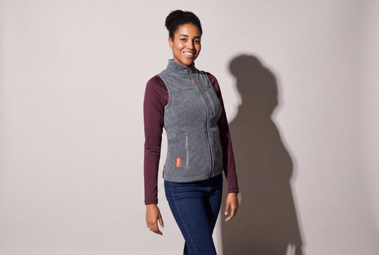 A woman wears a grey heated fleece vest zipped over a burgundy long-sleeve top and jeans, turned three quarters against a warm grey studio wall.