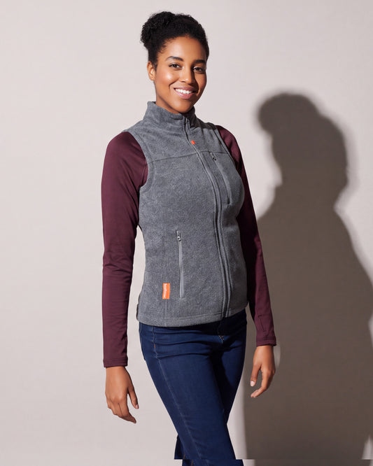 Une femme porte une veste chauffante en laine polaire grise par-dessus un chandail bordeaux à manches longues et des jeans, tournée de trois quarts devant un mur de studio gris chaud.