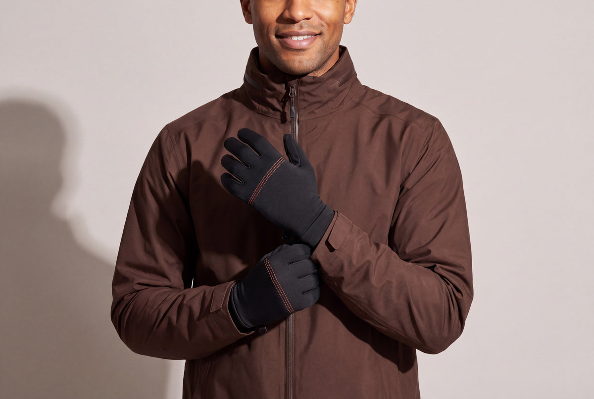 A man in a dark brown jacket rests his hands on his chest in thin black heated glove liners with orange stitching, against a warm grey studio wall, men’s frame