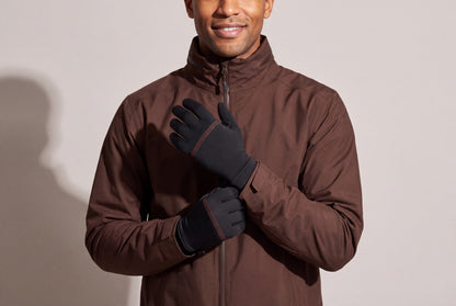 A man in a dark brown jacket rests his hands on his chest in thin black heated glove liners with orange stitching, against a warm grey studio wall, men’s frame