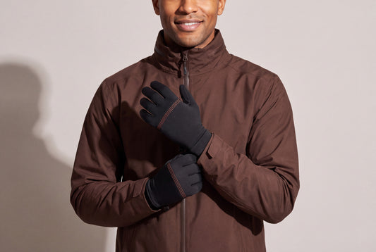 A man in a dark brown jacket rests his hands on his chest in thin black heated glove liners with orange stitching, against a warm grey studio wall, men’s frame