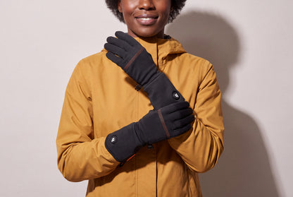 A woman in an ochre jacket crosses her hands in thin black heated glove liners with orange stitching, against a warm grey studio wall, women’s frame