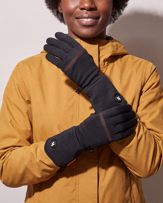 Une femme en manteau ocre croise les mains dans de minces sous-gants chauffants noirs à surpiqûres orange, devant un mur de studio gris chaud.