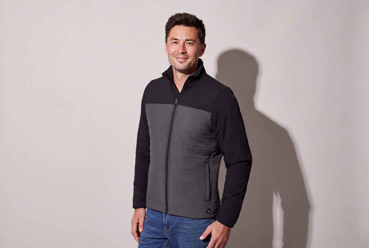 A man wears a black and charcoal heated puffer jacket zipped over jeans, standing three quarters against a warm grey studio wall.