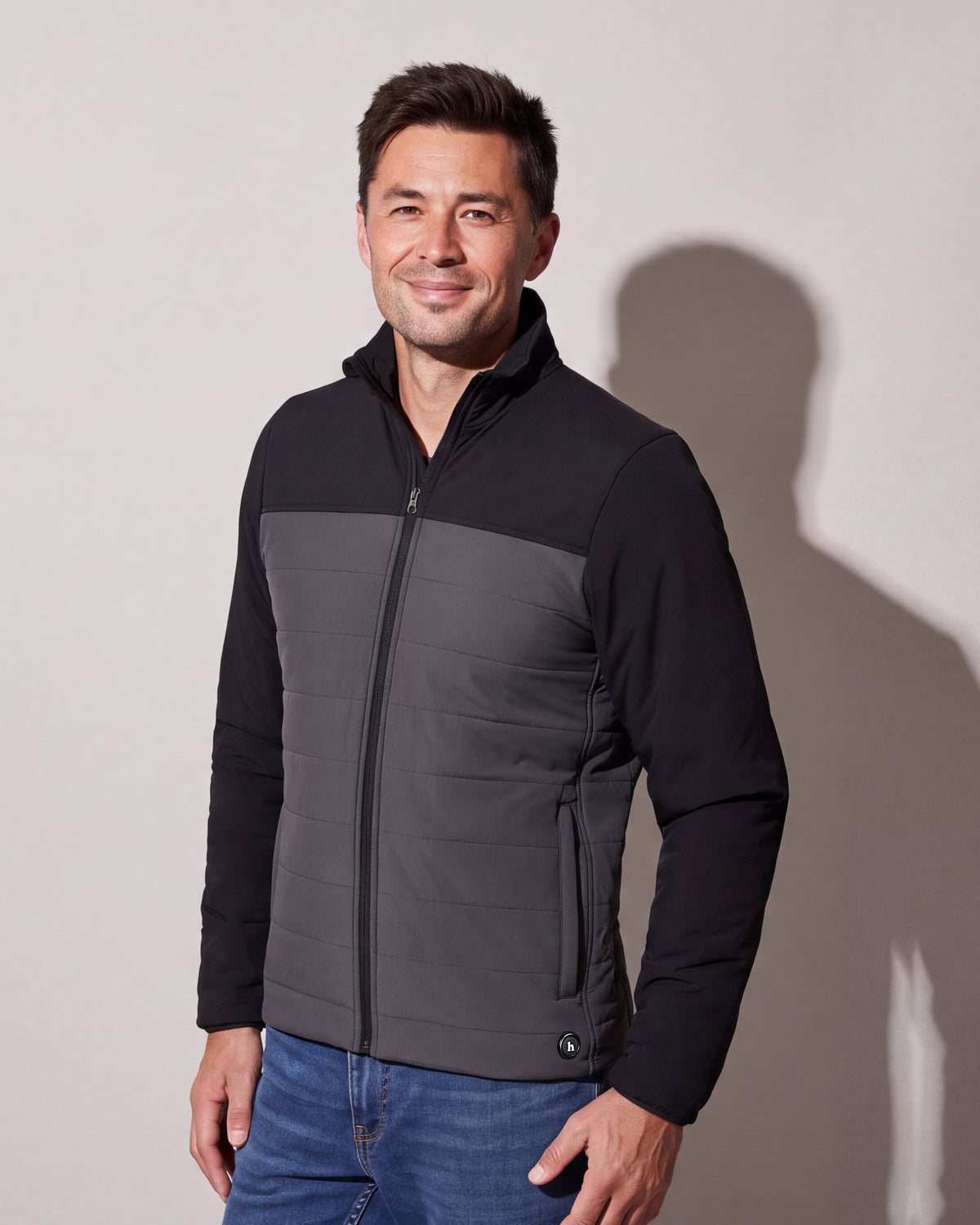 A man wears a black and charcoal heated puffer jacket zipped over jeans, standing three quarters against a warm grey studio wall.