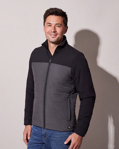 A man wears a black and charcoal heated puffer jacket zipped over jeans, standing three quarters against a warm grey studio wall.