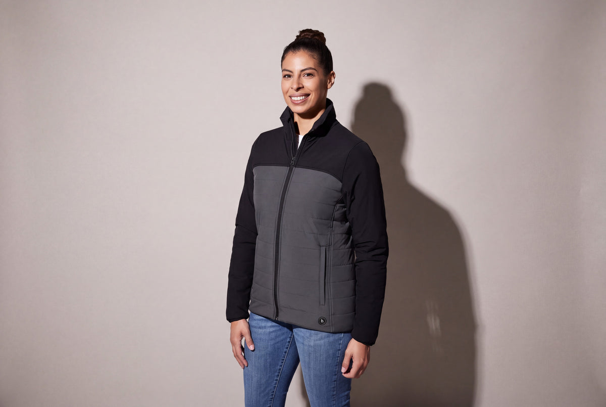 A woman wears a black and charcoal heated puffer jacket zipped over jeans, standing three quarters against a warm grey studio wall.