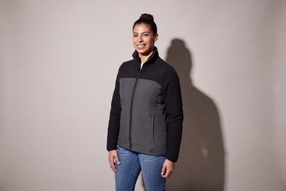 A woman wears a black and charcoal heated puffer jacket zipped over jeans, standing three quarters against a warm grey studio wall.