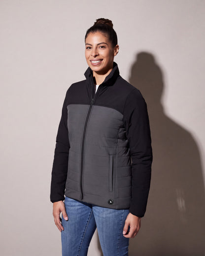 A woman wears a black and charcoal heated puffer jacket zipped over jeans, standing three quarters against a warm grey studio wall.