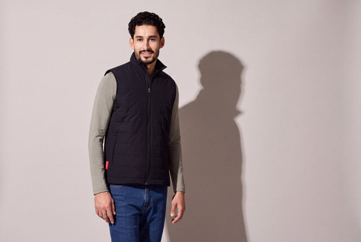 A man wears a black heated puffer vest with Bluetooth over a sage long-sleeve top and jeans, the red hem tab showing, against a warm grey studio wall, men’s frame