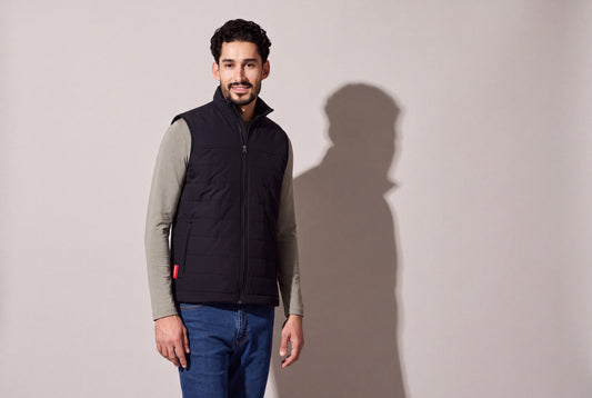 A man wears a black heated puffer vest with Bluetooth over a sage long-sleeve top and jeans, the red hem tab showing, against a warm grey studio wall, men’s frame