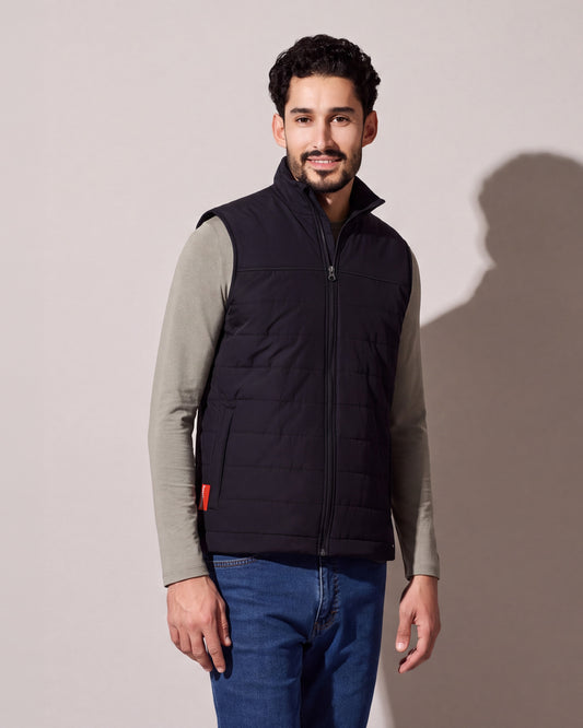 Un homme porte une veste chauffante matelassée noire avec Bluetooth par-dessus un chandail vert sauge à manches longues et des jeans, la languette rouge visible à l’ourlet, devant un mur de studio gris chaud.