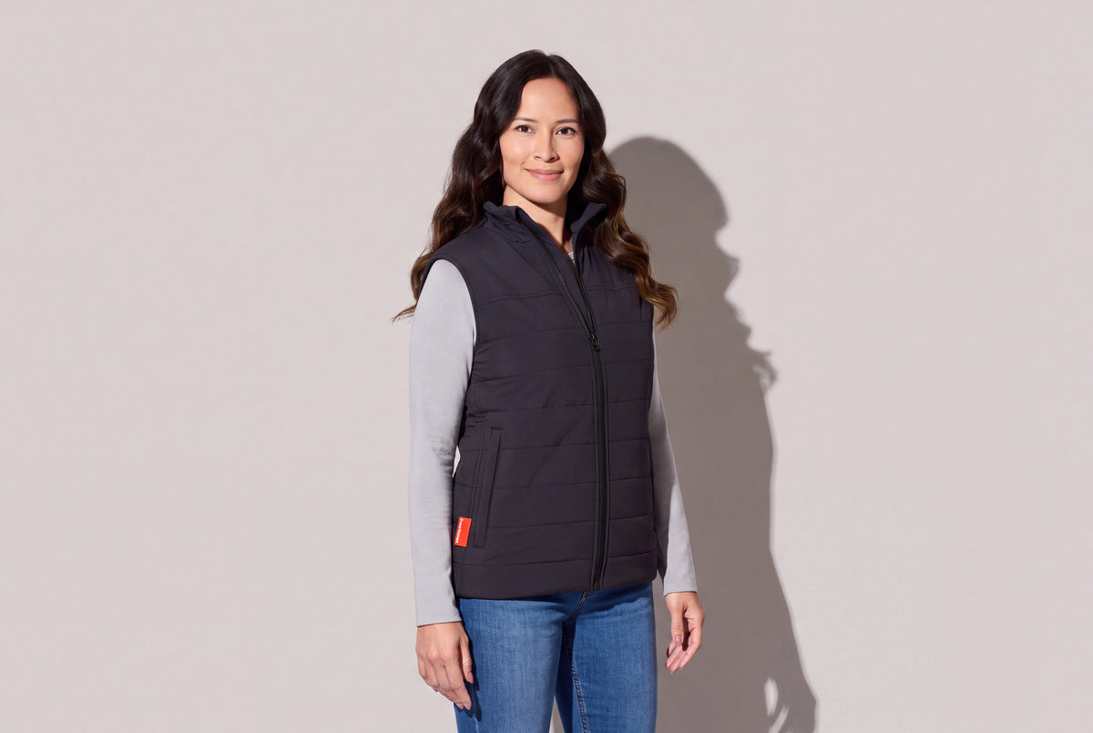 A woman wears a black heated puffer vest with Bluetooth over a grey long-sleeve top and jeans, the red hem tab showing, against a warm grey studio wall, women’s frame