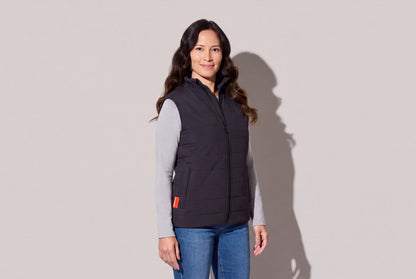 A woman wears a black heated puffer vest with Bluetooth over a grey long-sleeve top and jeans, the red hem tab showing, against a warm grey studio wall, women’s frame