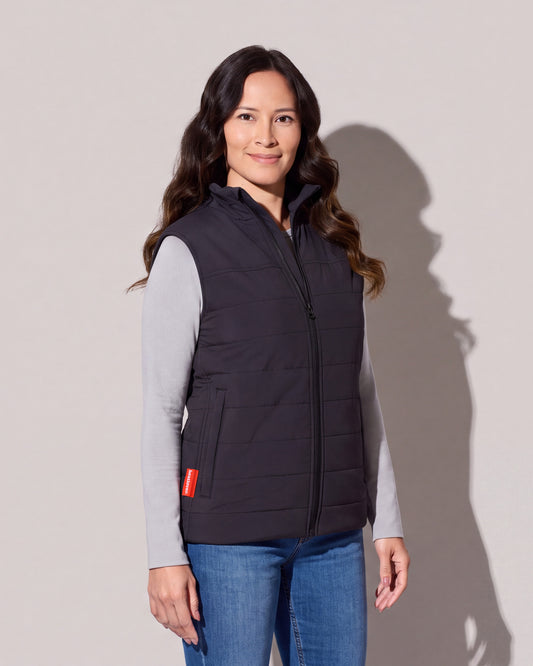 Une femme porte une veste chauffante matelassée noire avec Bluetooth par-dessus un chandail gris à manches longues et des jeans, la languette rouge visible à l’ourlet, devant un mur de studio gris chaud.