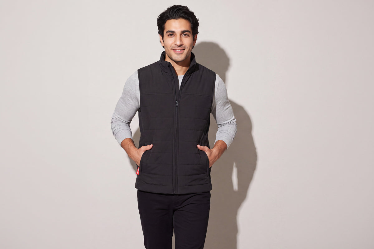 A man wears a black heated puffer vest over a grey long-sleeve top, both hands in the pockets, against a warm grey studio wall, men’s frame