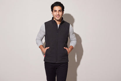 A man wears a black heated puffer vest over a grey long-sleeve top, both hands in the pockets, against a warm grey studio wall, men’s frame