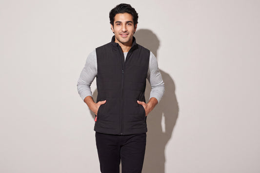 A man wears a black heated puffer vest over a grey long-sleeve top, both hands in the pockets, against a warm grey studio wall, men’s frame