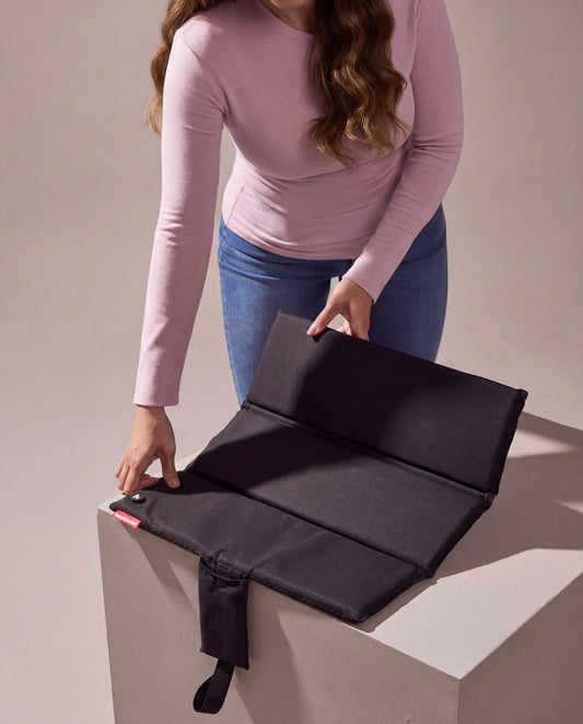 A woman in a pink long-sleeve top unfolds the black heated seat cushion on a pale plinth against a warm grey studio wall.