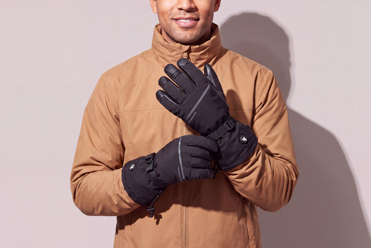 A man in a tan winter jacket holds his black heated gloves together at chest height against a warm grey studio wall, men’s frame