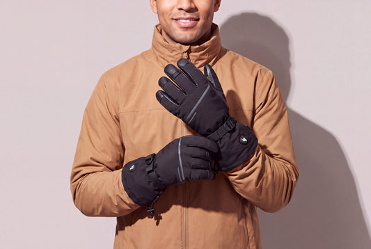 A man in a tan winter jacket holds his black heated gloves together at chest height against a warm grey studio wall, men’s frame