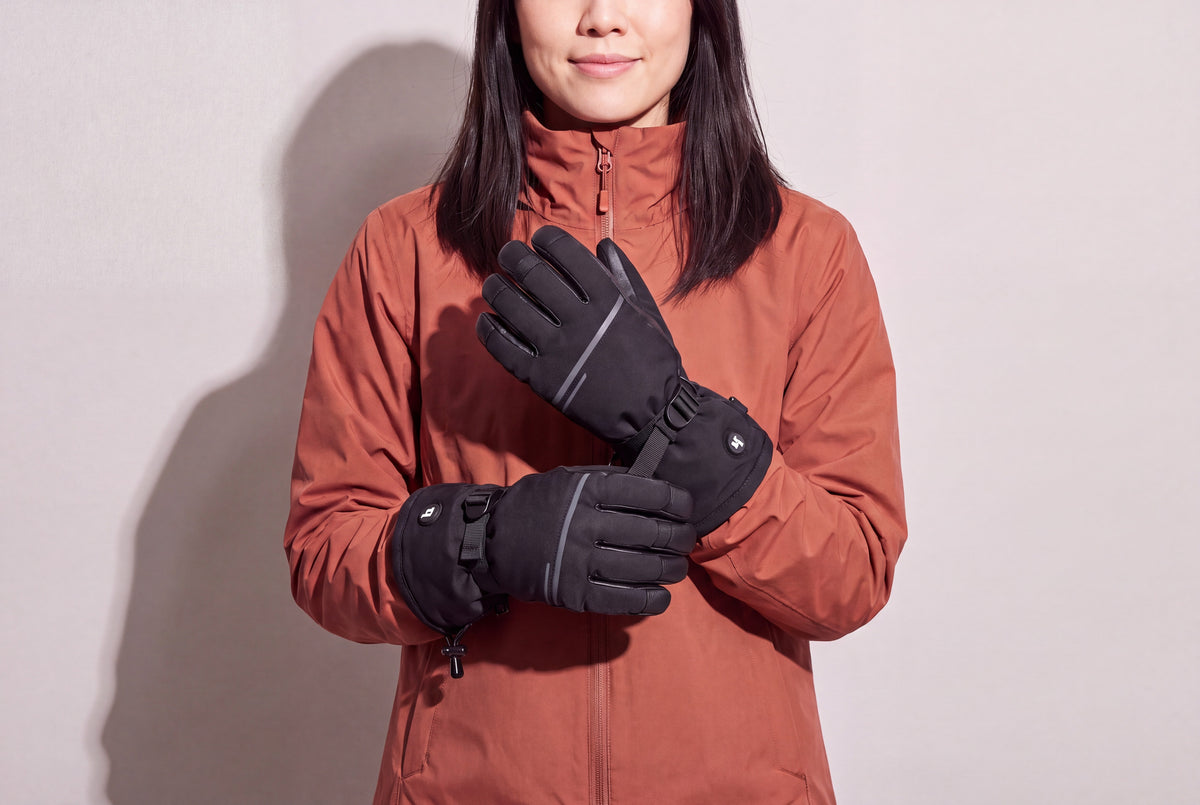 A woman in a rust winter jacket holds her black heated gloves crossed at chest height against a warm grey studio wall, women’s frame