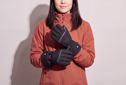 A woman in a rust winter jacket holds her black heated gloves crossed at chest height against a warm grey studio wall, women’s frame