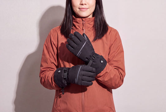 A woman in a rust winter jacket holds her black heated gloves crossed at chest height against a warm grey studio wall, women’s frame
