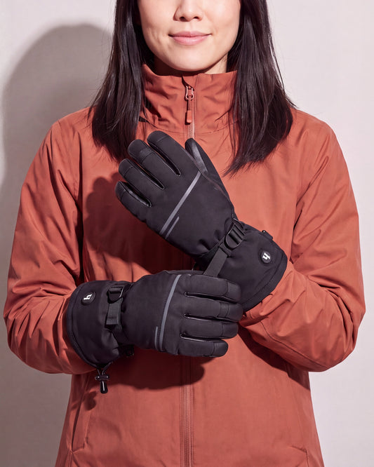 Une femme en manteau d’hiver rouille tient ses gants chauffants noirs croisés à hauteur de poitrine devant un mur de studio gris chaud.