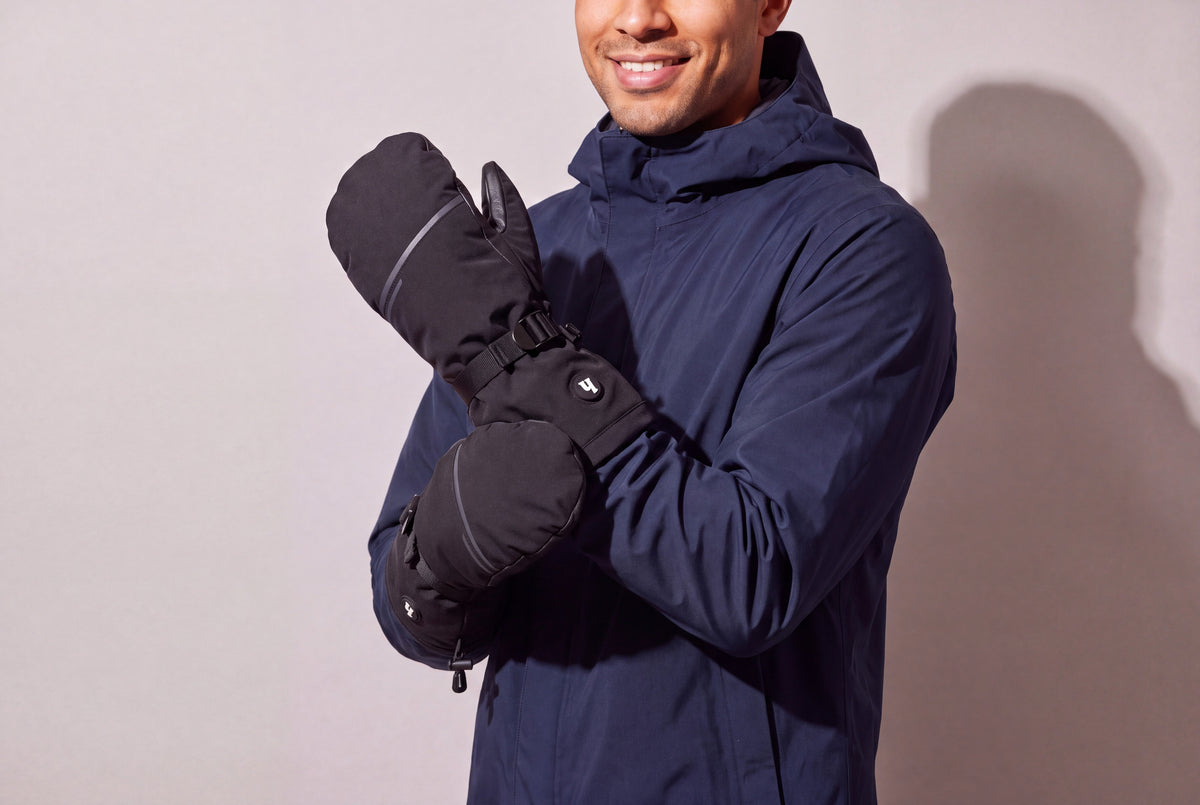 A man in a navy hooded shell jacket crosses his arms wearing black heated mitts, seen three quarters against a warm grey studio wall, men’s frame