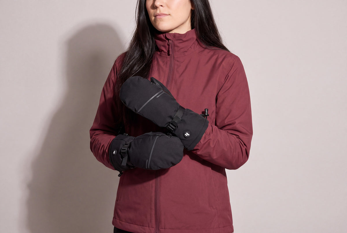 A woman in a burgundy shell jacket holds her black heated mitts together at chest height against a warm grey studio wall, women’s frame