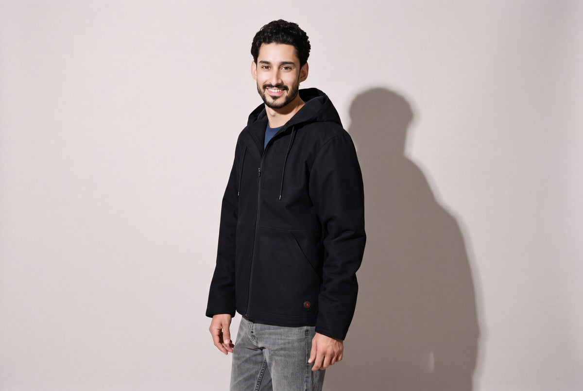 A man wears a navy hooded heated canvas work jacket over grey jeans, standing three quarters against a warm grey studio wall.