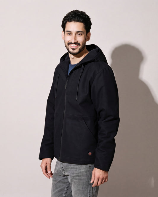Un homme porte un manteau de travail chauffant en toile noire à capuchon par-dessus des jeans gris, debout de trois quarts devant un mur de studio gris chaud.