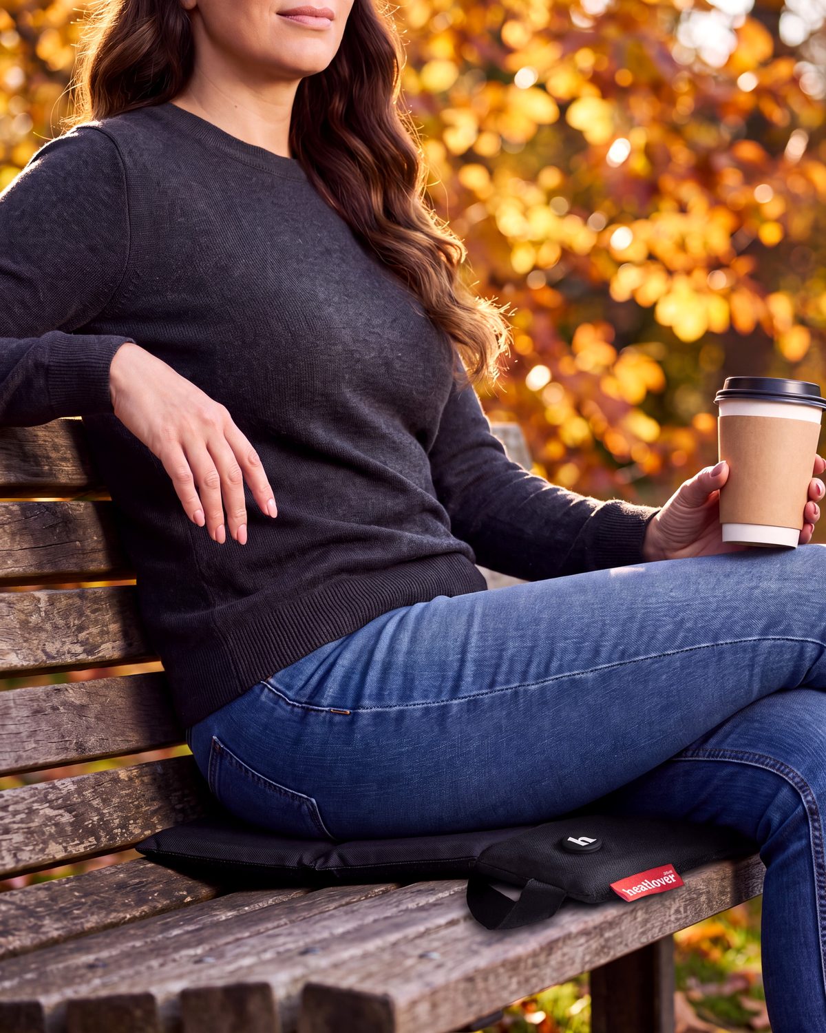 Une femme assise sur un banc de parc en automne, sur un coussin chauffant heatlover noir, un café à la main.