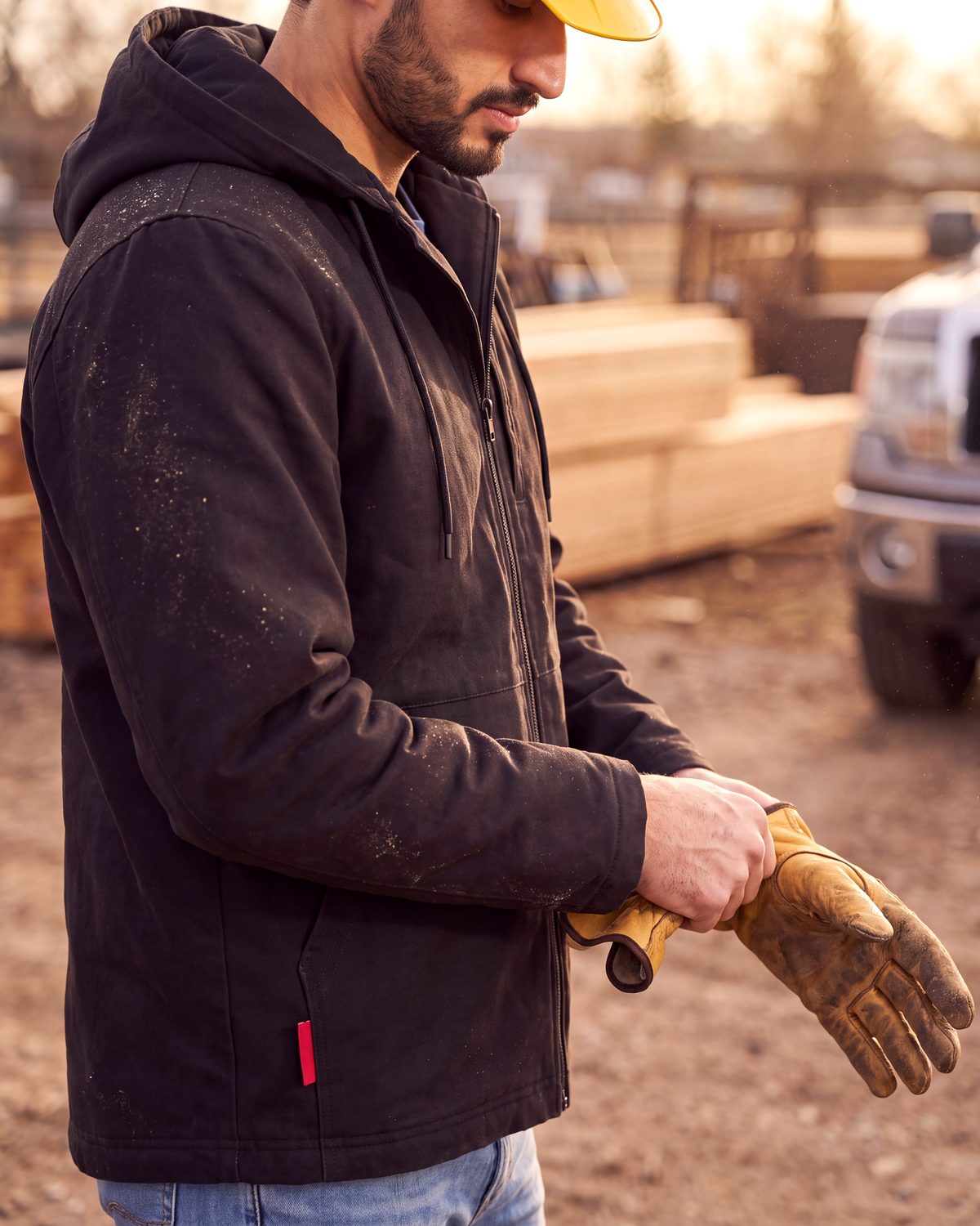 Un travailleur en manteau de toile heatlover noir et casque de chantier enfile un gant de travail en cuir.